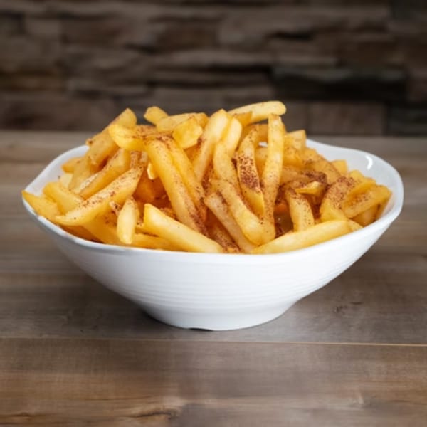 A white bowl filled with golden french fries, lightly seasoned with dark spices, on a wooden table.