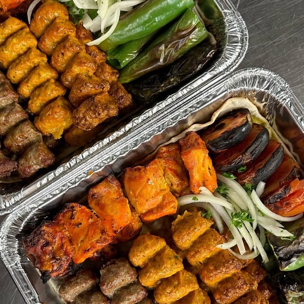 Two foil containers filled with various grilled Mediterranean kabobs, vegetables, and onions.